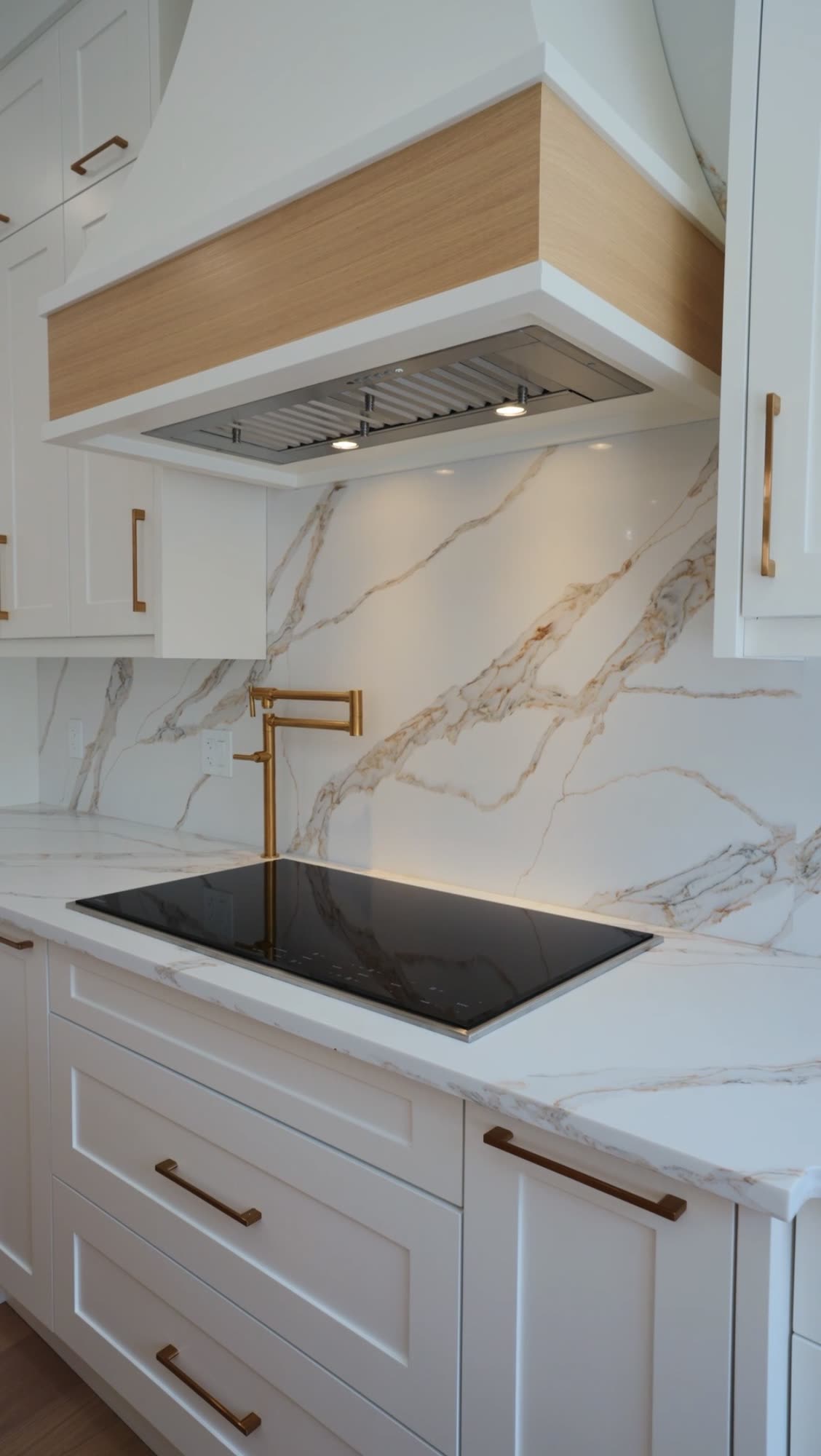 Cooktop below an oak banded hood with a brass pot filler on the marble wall